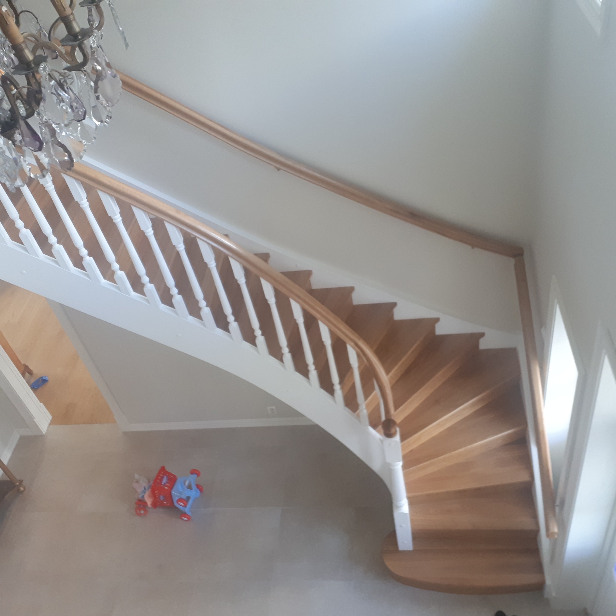 Klatka schodowa z drewnianymi stopniami i białymi słupkami balustrady, widok z góry. W tle widoczny żyrandol i dziecięca zabawka.