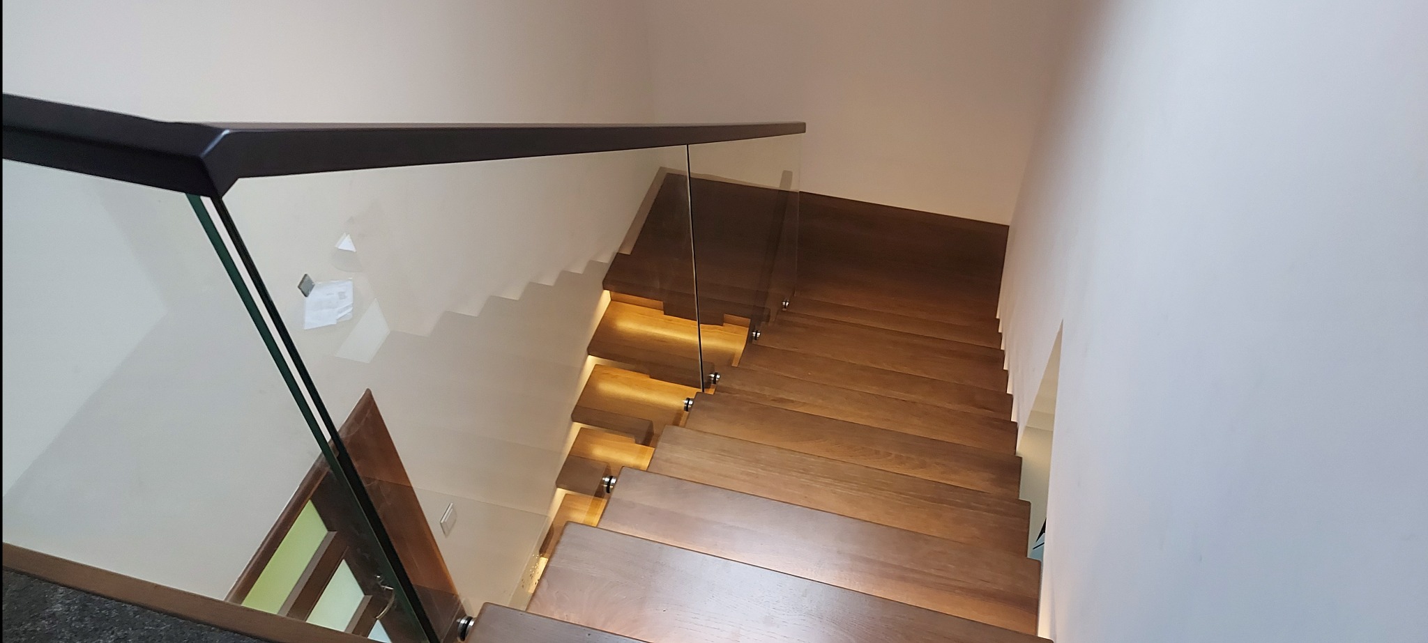 Nowoczesne schody z drewna, z podświetleniem stopni i szklaną barierką. Widok z góry na jasne stopnie.
