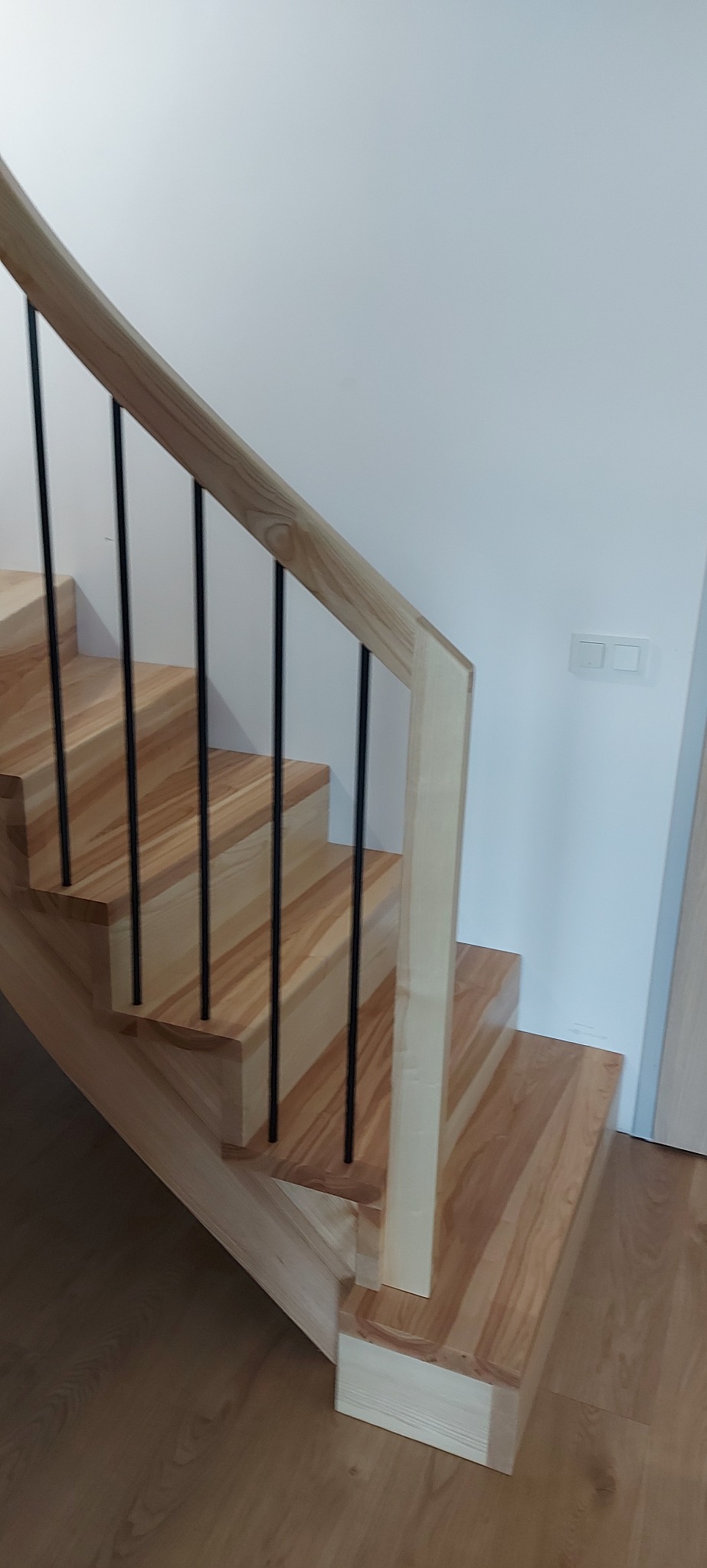 Jasne, drewniane schody z czarnymi, metalowymi prętami. Naturalne wykończenie drewna, minimalistyczny design, widoczne detale łączeń. Oświetlenie dzienne, wnętrze domu.