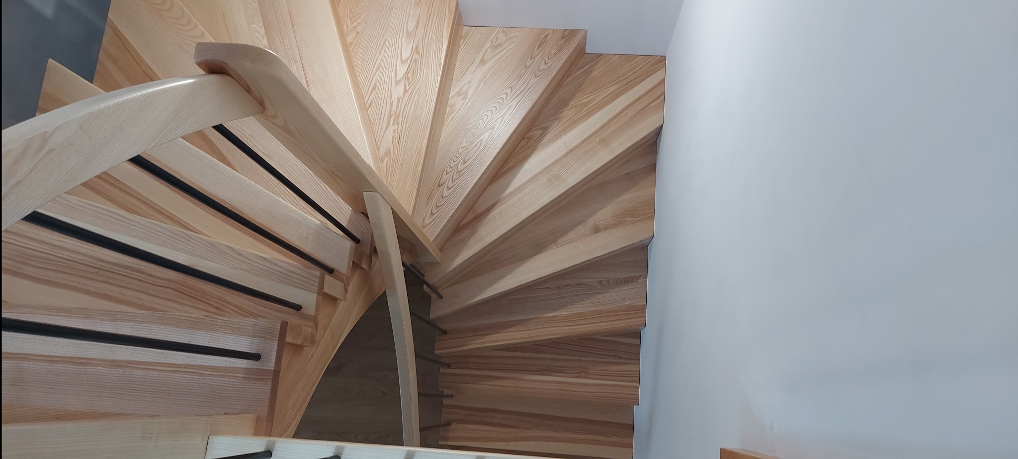 Spiralne schody z jasnego drewna z czarnymi, metalowymi prętami w balustradzie, widziane z góry, z fragmentem białej ściany po prawej stronie.