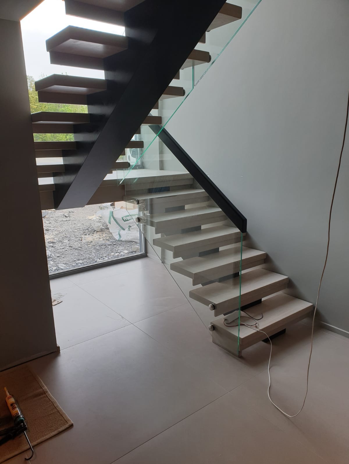 Nowoczesne schody wspornikowe z drewnianymi stopniami, czarną konstrukcją i szklaną balustradą, widoczne z dołu, z oknem w tle i narzędziami na podłodze.