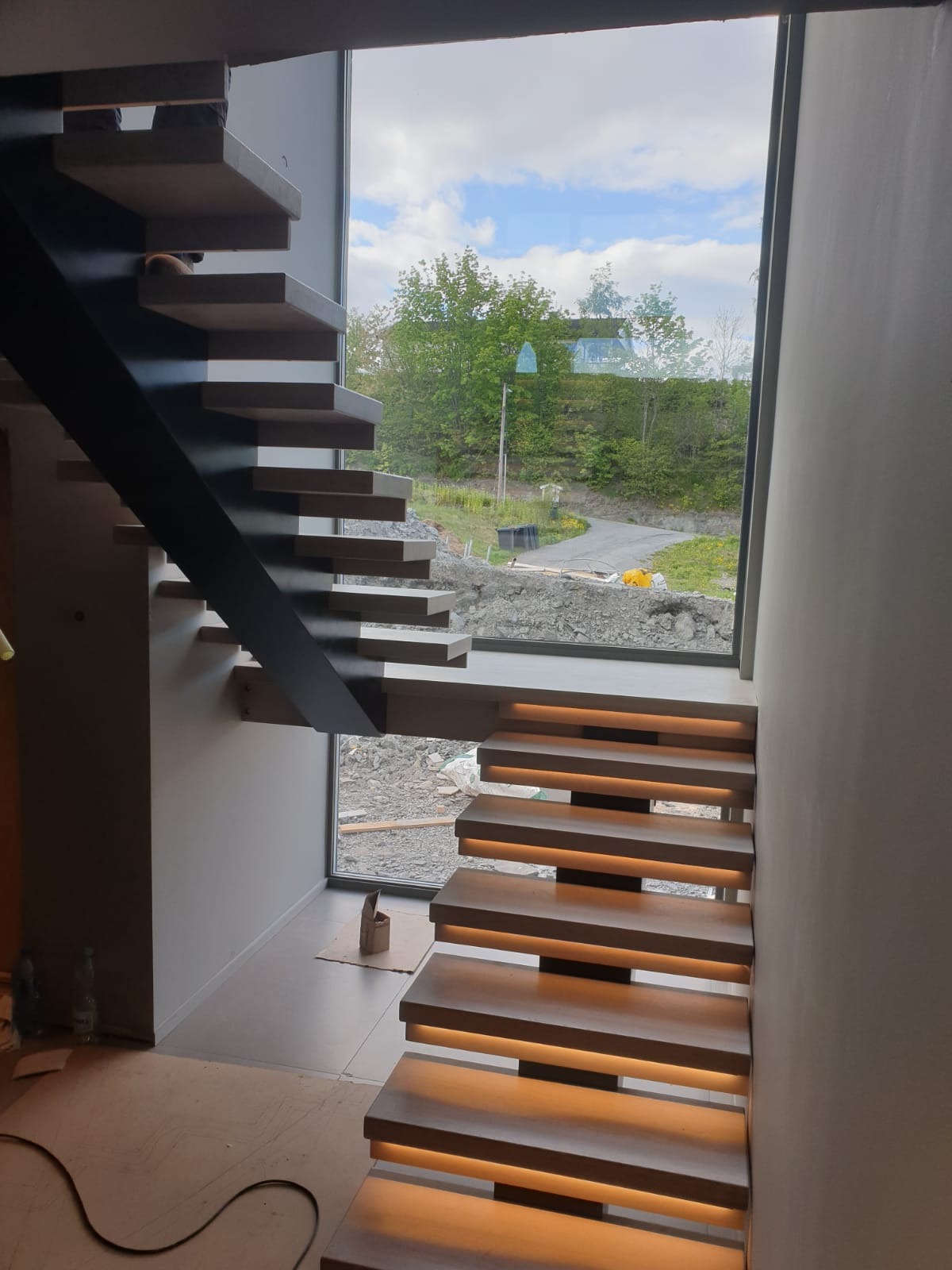 Nowoczesne schody z podświetleniem LED, wspornikowa konstrukcja z czarnym elementem nośnym, widok na zewnątrz przez duże okno. Detal architektoniczny wnętrza.