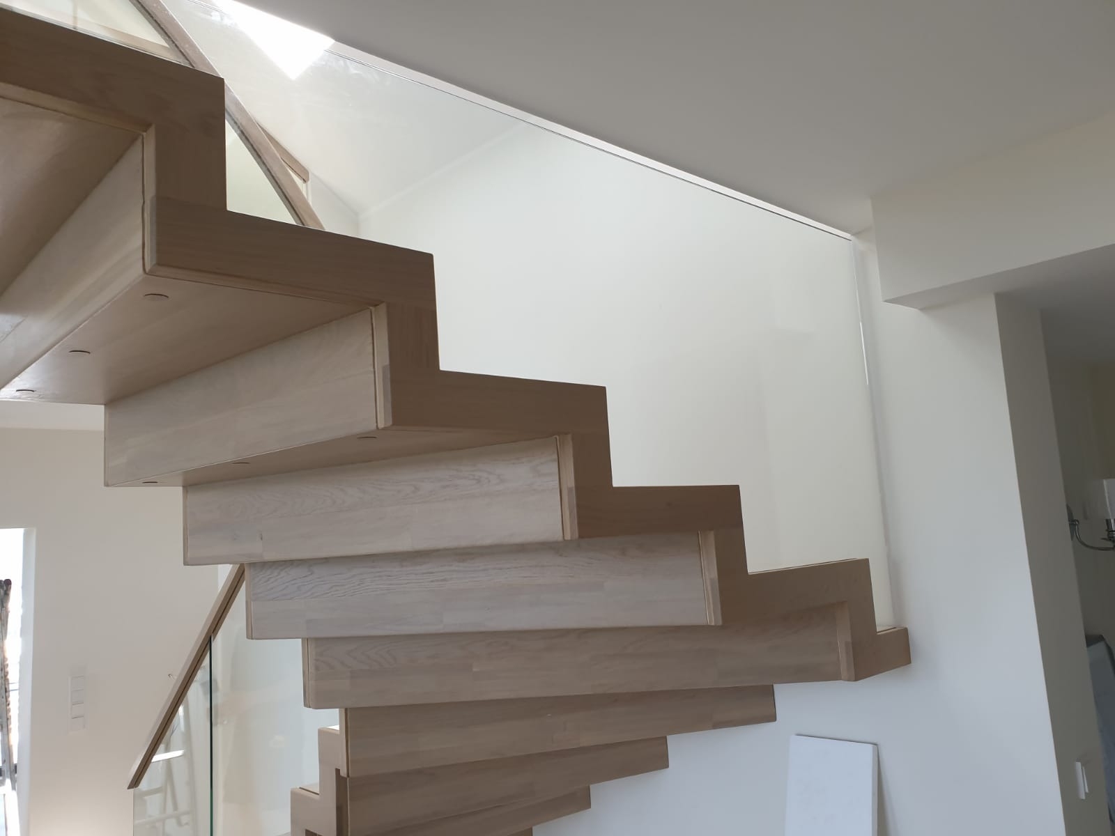 Nowoczesne schody z jasnego drewna, bezstopniowe, z szklaną balustradą, widok z dołu, minimalistyczny design, dobrze oświetlone wnętrze.