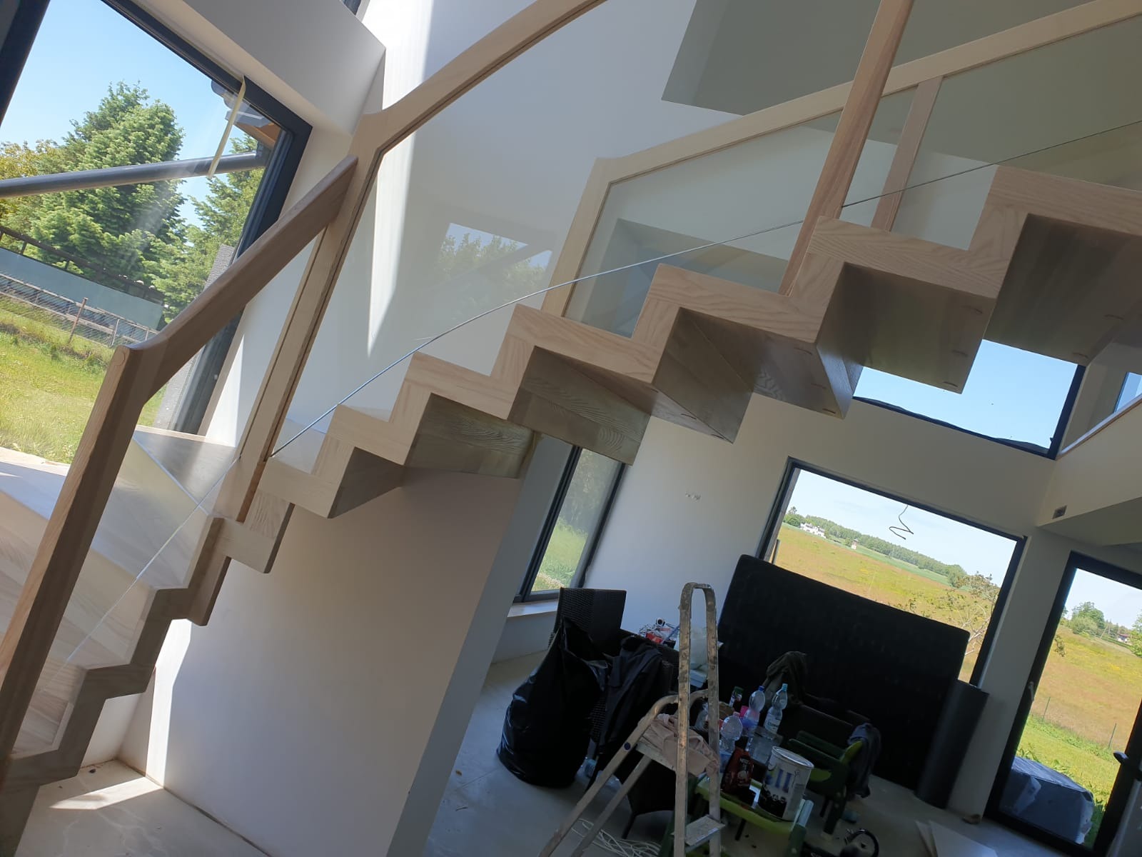 Nowoczesne schody z jasnego drewna z szklaną balustradą w przestronnym wnętrzu z dużymi oknami. Widoczne elementy wykończeniowe i narzędzia.