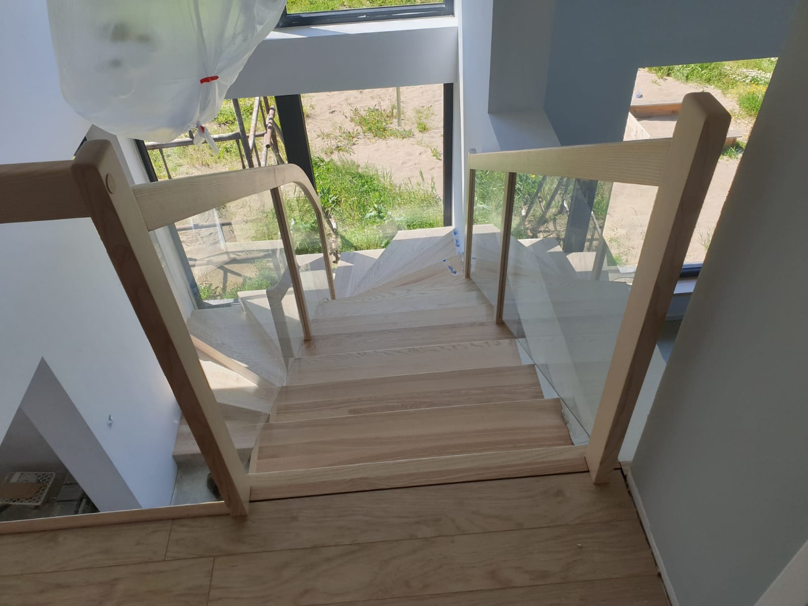Jasne schody z drewna z przezroczystymi szklanymi panelami. Widok z góry na zakręcające schody w nowoczesnym wnętrzu z dużym oknem.
