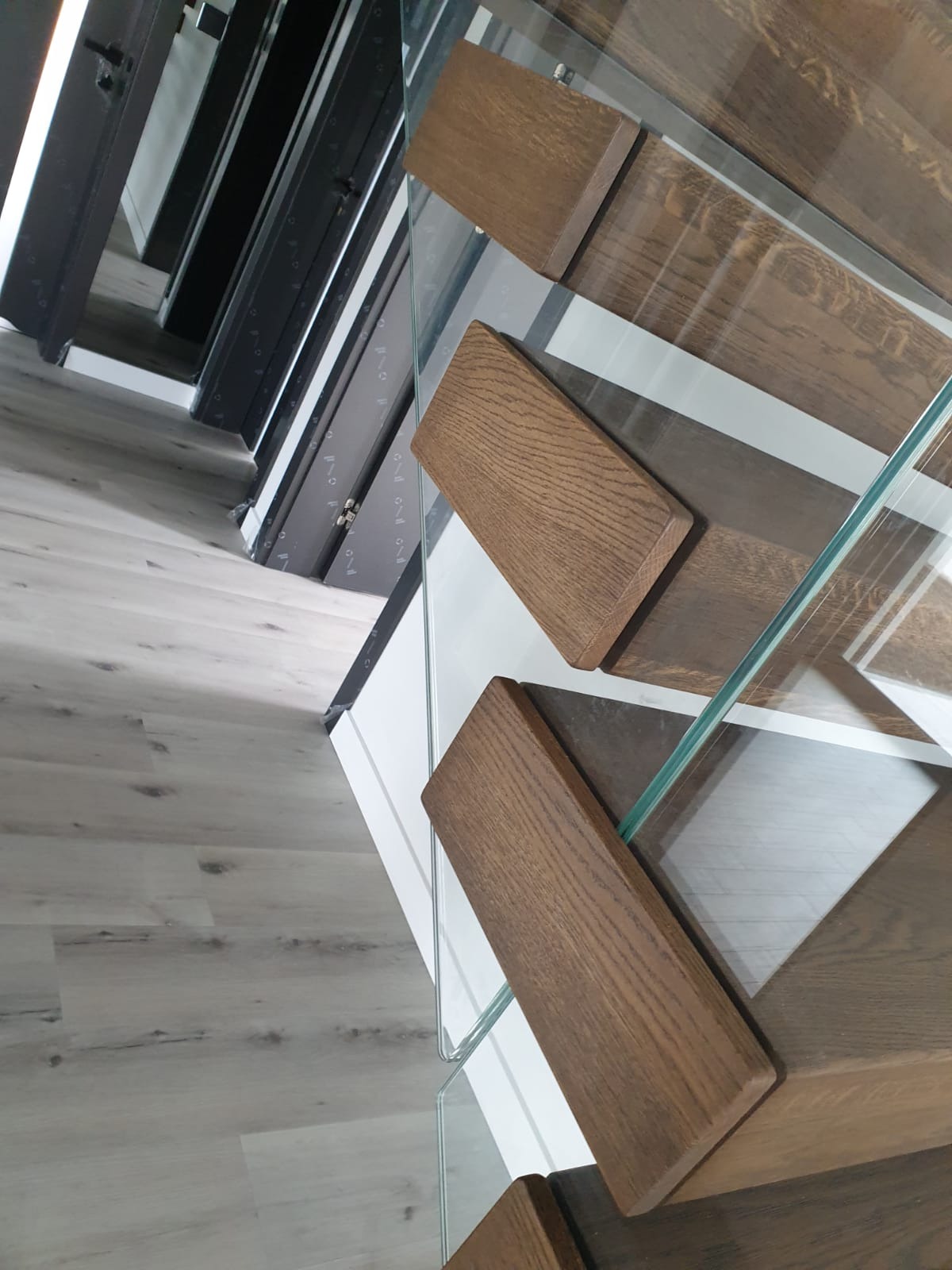 Nowoczesne schody z drewnianymi stopniami i szklaną balustradą, widoczne z góry, z podłogą imitującą drewno w tle.