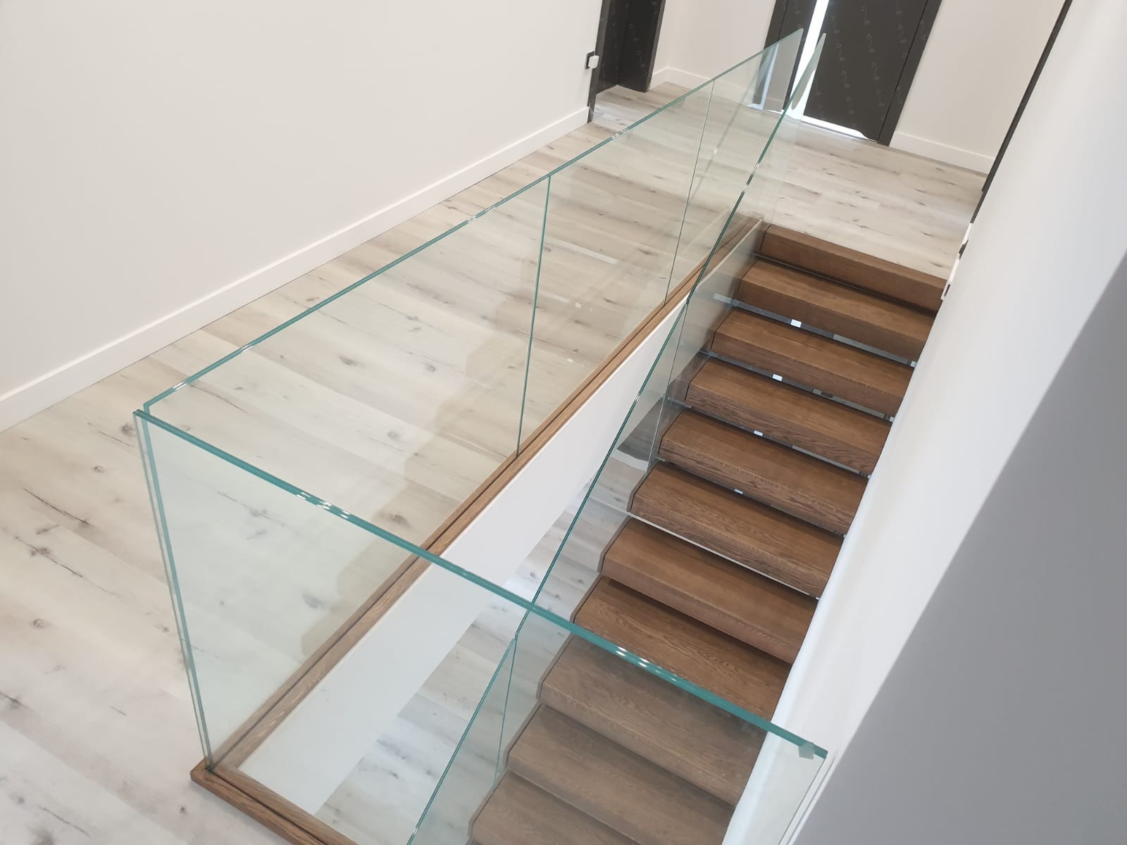 Nowoczesne schody z drewnianymi stopniami i szklaną balustradą, widok z góry, jasne wnętrze z parkietem i białymi ścianami, czarne drzwi w tle.