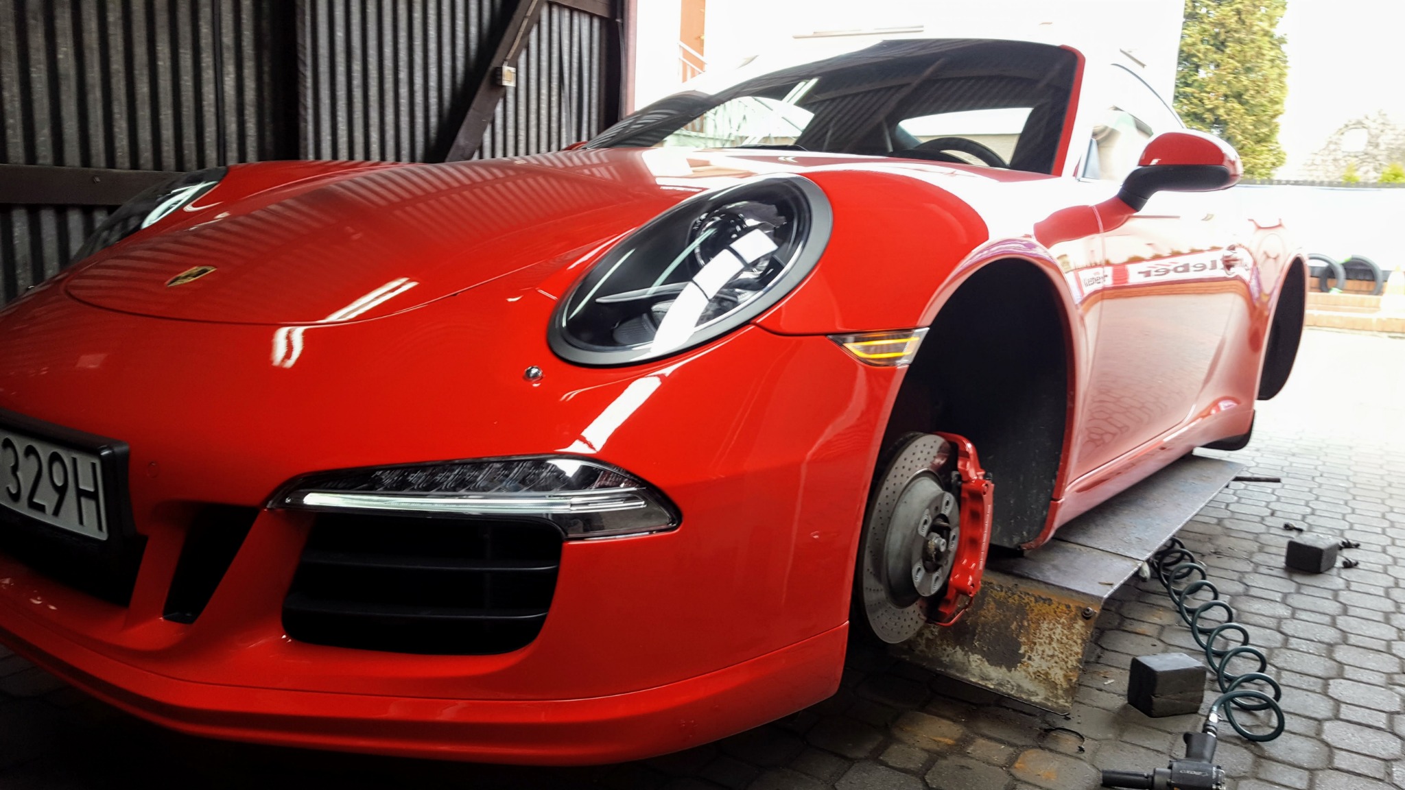 Czerwone Porsche 911 na podnośniku warsztatowym, bez koła, widoczne tarcze hamulcowe i narzędzia pneumatyczne, wnętrze warsztatu.