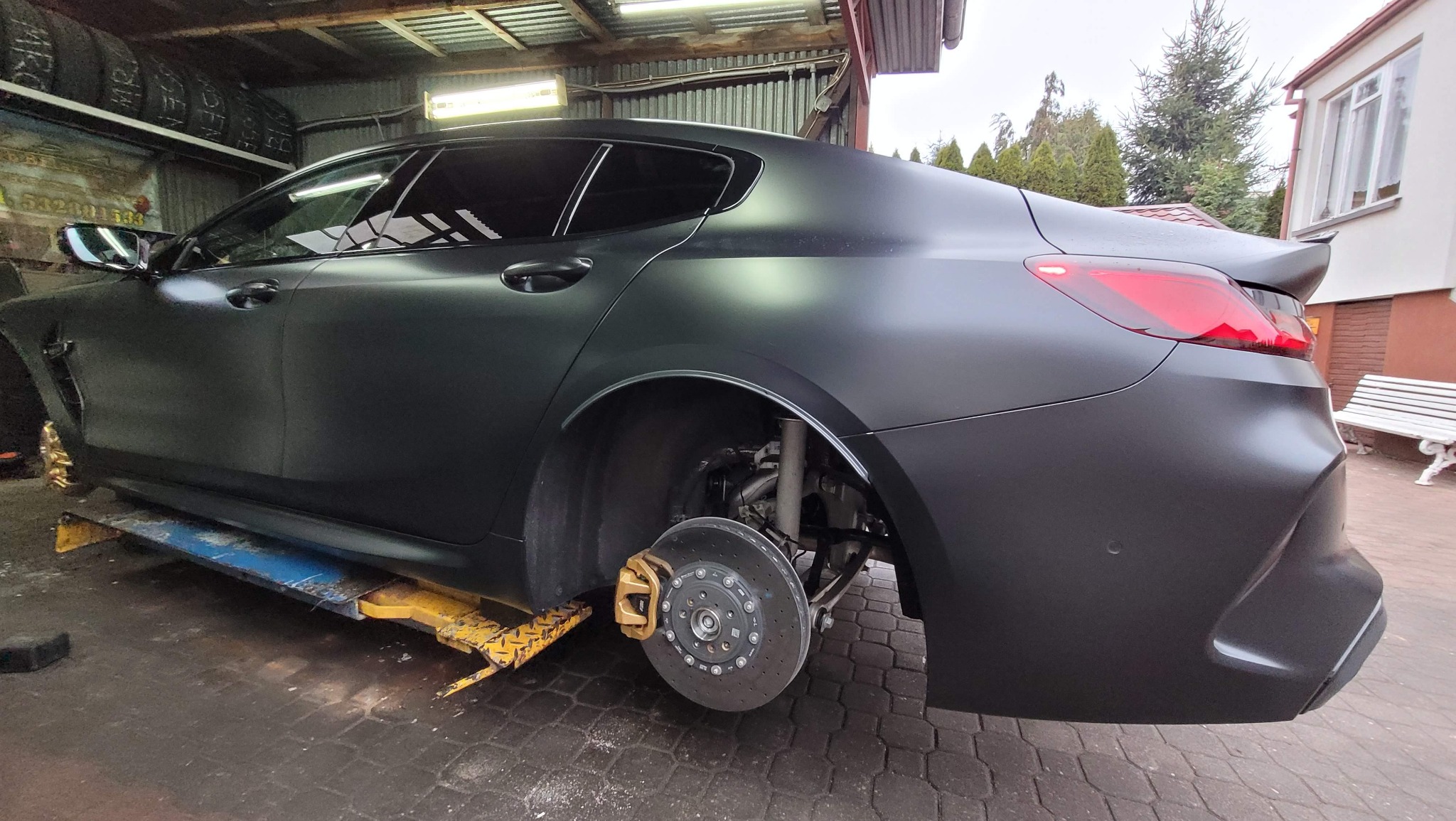 Matowy czarny samochód na podnośniku w warsztacie, brak koła, widoczne złote zaciski hamulcowe i tarcza hamulcowa.