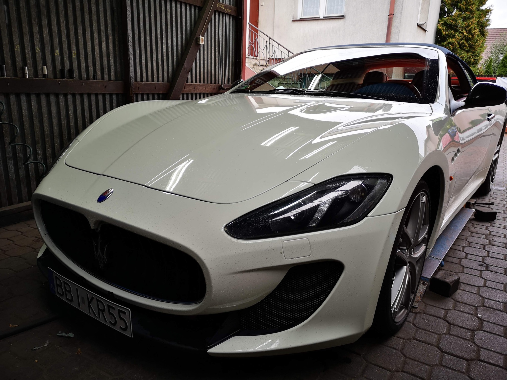 Białe Maserati GranTurismo na podnośniku w warsztacie, widoczne krople wody na karoserii po umyciu, z detalami wnętrza w odcieniach brązu widocznymi przez otwarte okno.