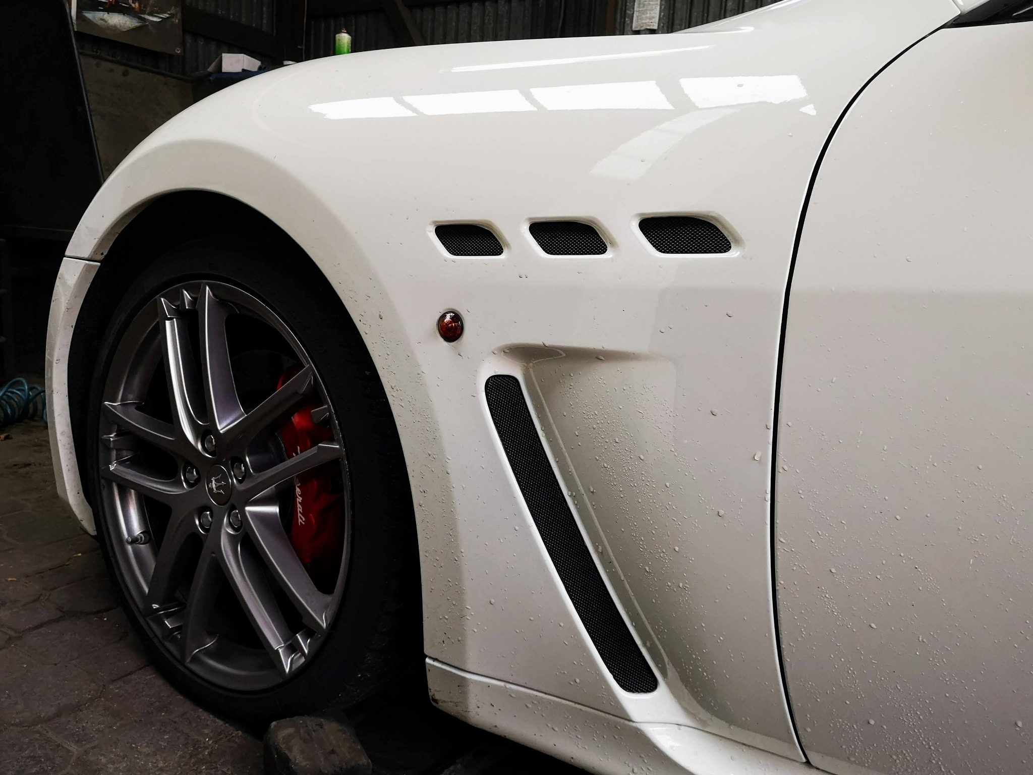 Biały samochód marki Maserati z widocznymi wlotami powietrza na błotniku i czerwonymi zaciskami hamulcowymi za szarą felgą.