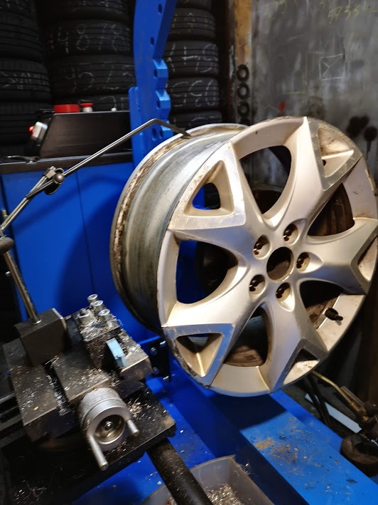 Aluminiowa felga na wyważarce z widocznymi opiłkami metalu, w tle stos opon i ściana z notatkami.