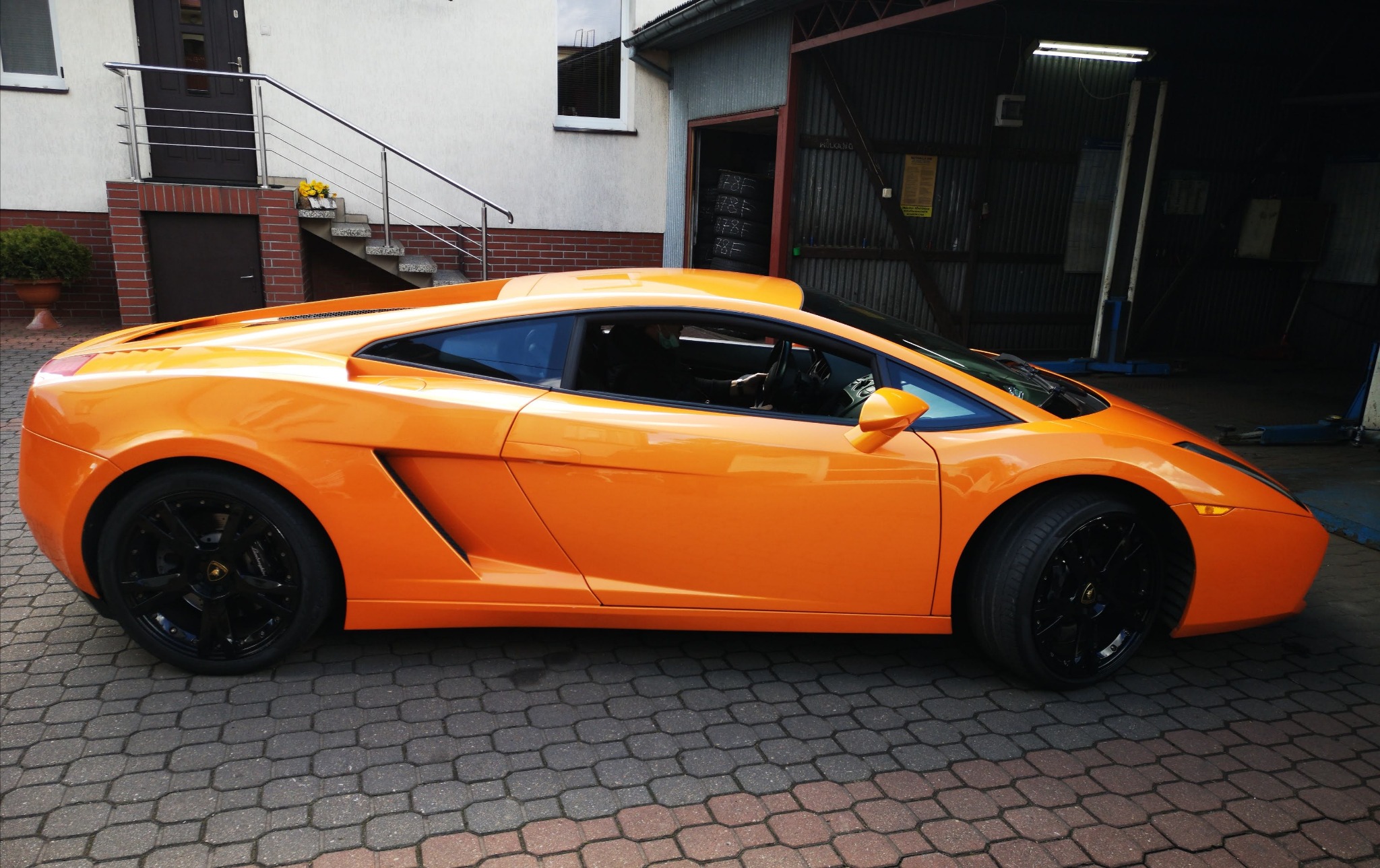 Pomarańczowe Lamborghini Gallardo zaparkowane przed warsztatem samochodowym z widocznymi oponami w tle.