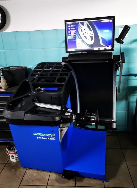 Niebieskie urządzenie Hofmann geodyna 8200p do wyważania kół samochodowych z monitorem wyświetlającym obraz koła, stojące w warsztacie z kafelkami na ścianie.