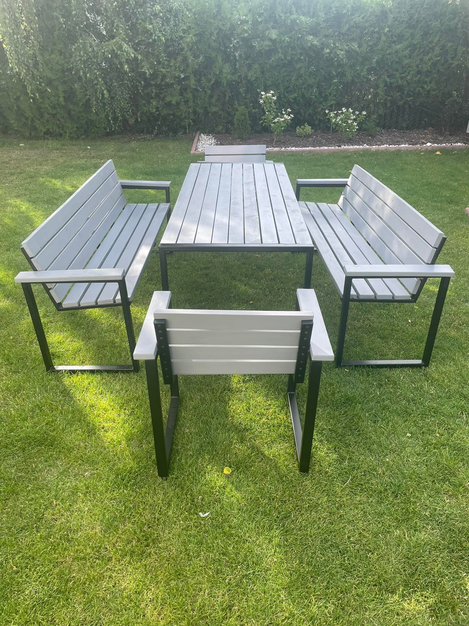 Zestaw mebli ogrodowych: stół z blatem z desek w odcieniach szarości i czarnych metalowych nogach, otoczony dwiema ławkami i krzesłem w tym samym stylu, ustawiony na trawniku przed żywopłotem.