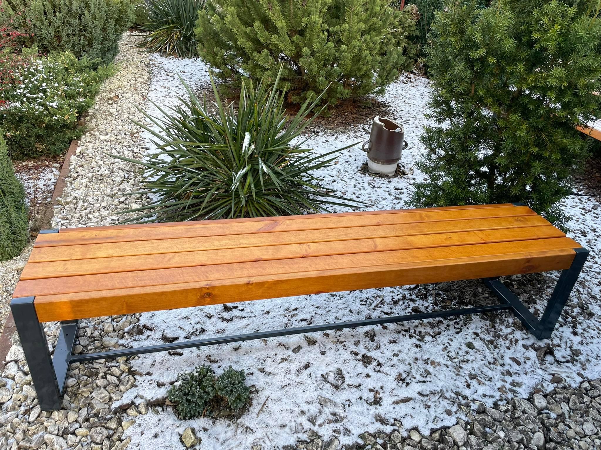 Drewniana ławka ogrodowa z metalowymi nogami, ustawiona na żwirowym podłożu z delikatnym śniegiem, otoczona zielenią w zimowej scenerii.