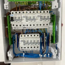 Otwarta skrzynka rozdzielcza z bezpiecznikami ETI i Eaton, widoczne przewody w różnych kolorach, listwy zaciskowe i elementy montażowe wewnątrz białej obudowy.