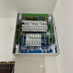 Valentyn Lohvynenko - Otwarta skrzynka elektryczna ETI IZ12/IF/12 z widocznymi bezpiecznikami, przewodami w kolorach niebieskim, brązowym i zielono-żółtym, zamontowana na białej ścianie z widocznymi śladami po pracach...