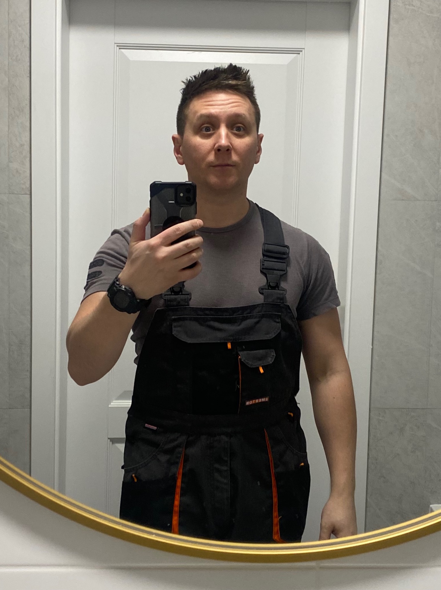 Mężczyzna w roboczym kombinezonie pozuje do selfie w łazience z białymi drzwiami i szarymi płytkami, widoczny w lustrze z pozłacaną ramą.