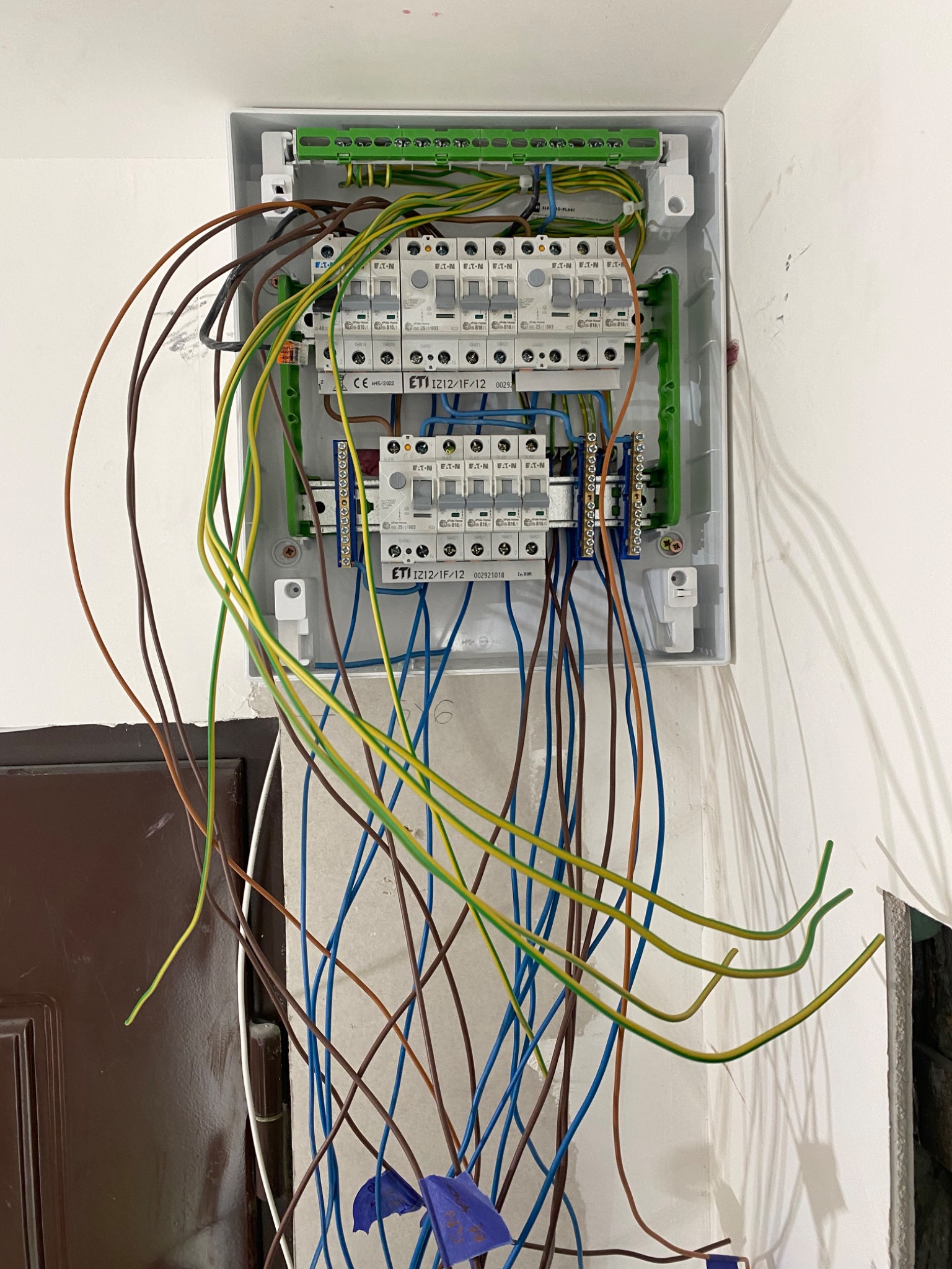 Otwarta skrzynka rozdzielcza z widocznymi bezpiecznikami ETI i licznymi przewodami o różnych kolorach, częściowo zwisającymi luźno, w trakcie podłączania wewnątrz budynku.