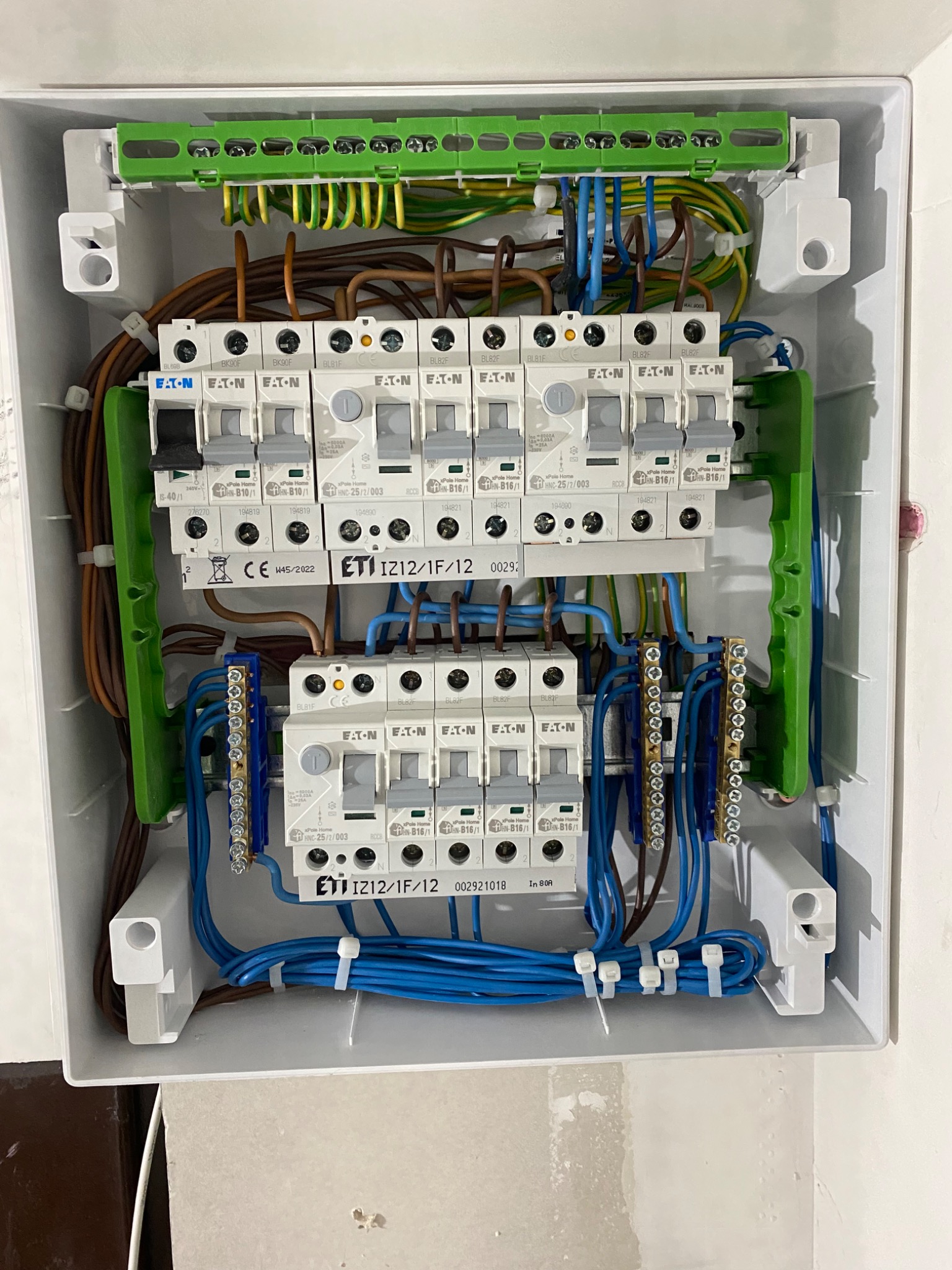 Otwarta skrzynka rozdzielcza z bezpiecznikami ETI i Eaton, widoczne przewody w różnych kolorach, listwy zaciskowe i elementy montażowe wewnątrz białej obudowy.