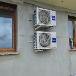 Dwa białe klimatyzatory Haier zamontowane jeden nad drugim na szarej, surowej ścianie budynku, widoczne okna z brązowymi ramami i pianka montażowa na parapecie.