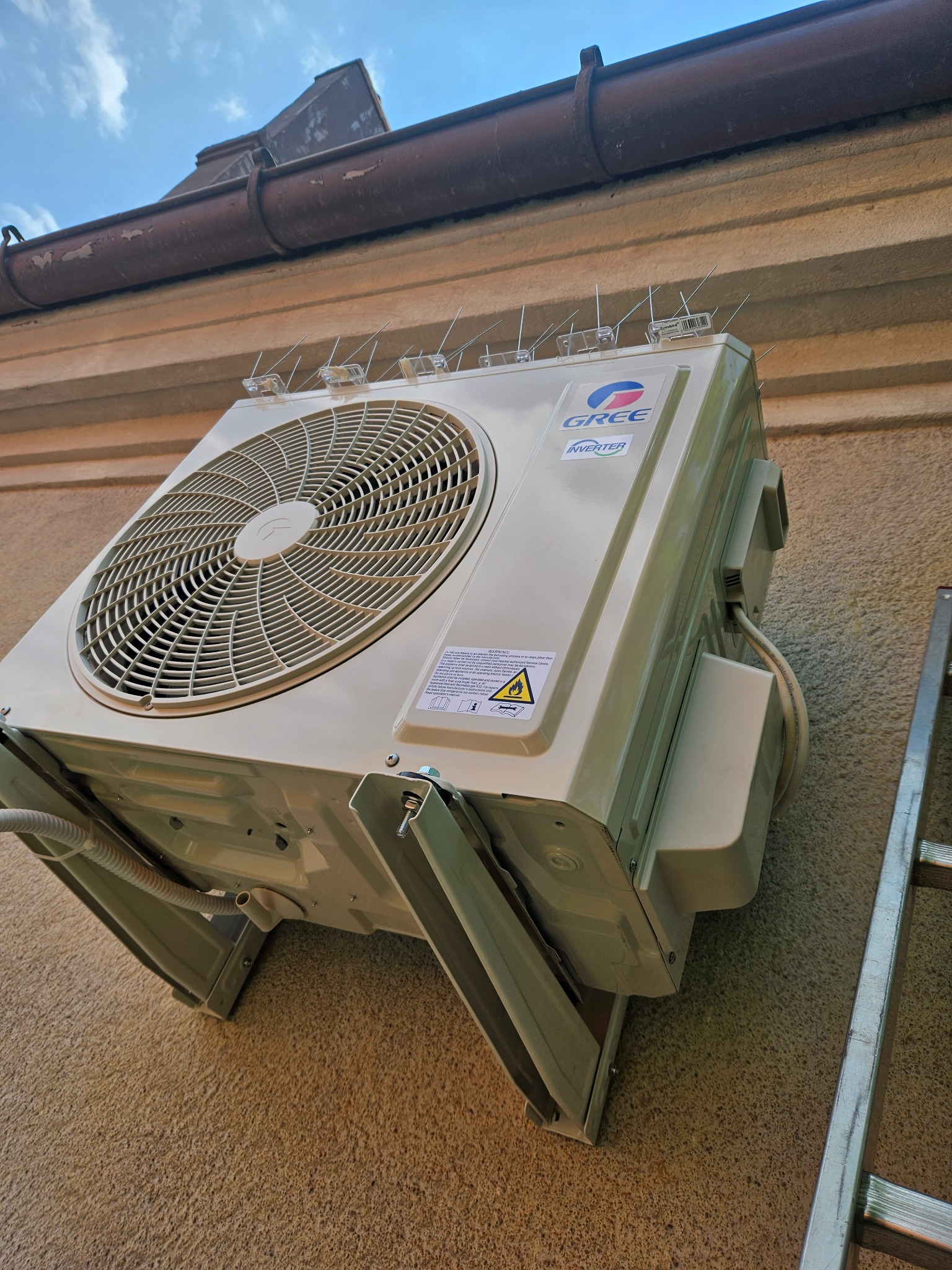 Zewnętrzna jednostka klimatyzacji marki Gree zamontowana na elewacji budynku, zabezpieczona kolcami przeciw ptakom, widoczna drabina oparta o ścianę.