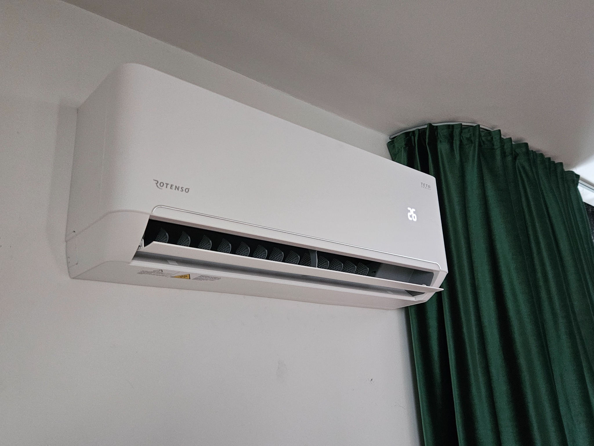 Biała klimatyzacja Rotenso zamontowana na ścianie, widoczny wyświetlacz z temperaturą 26 stopni, obok zielone, welurowe zasłony.