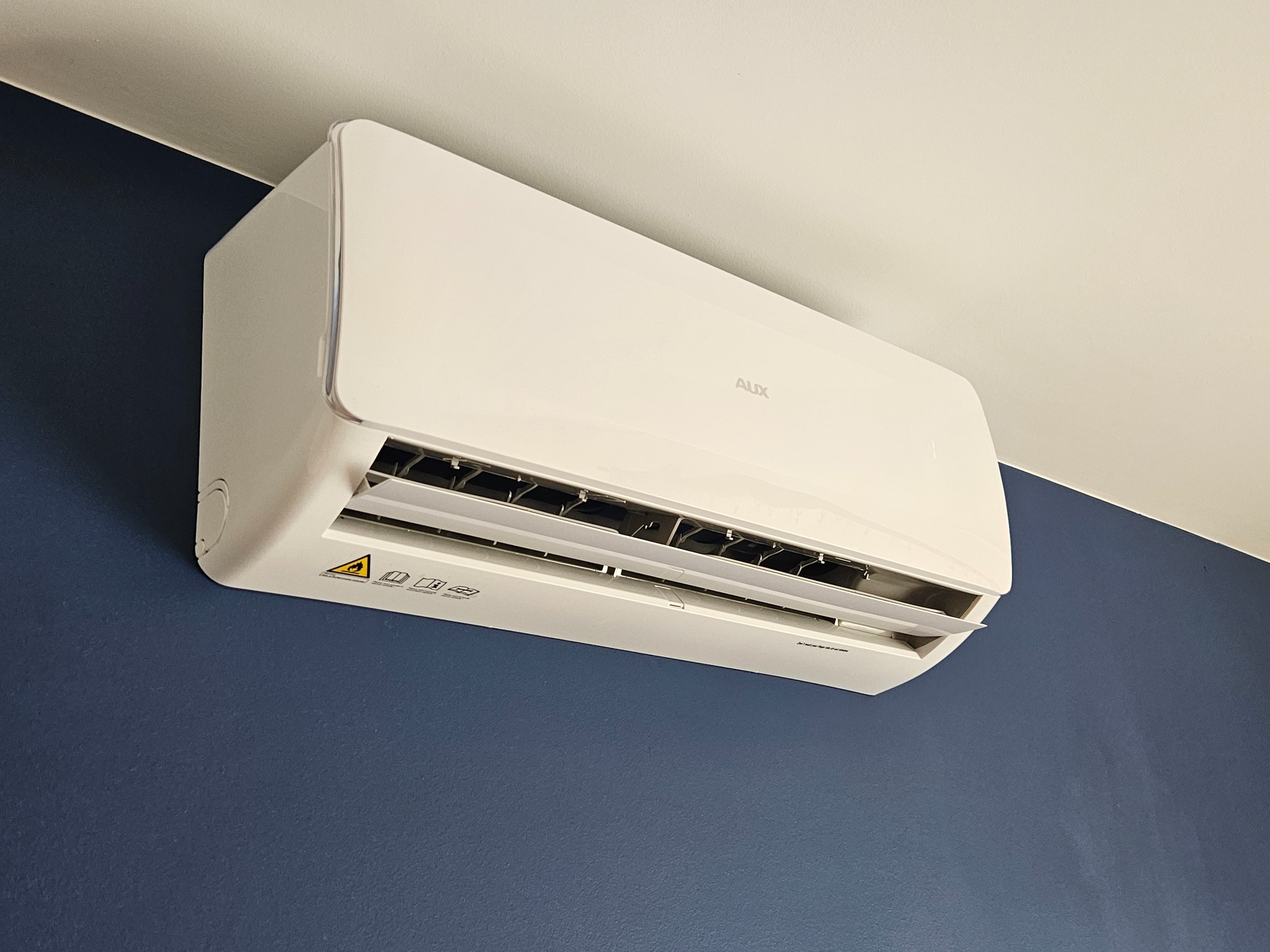 Biała klimatyzacja marki AUX zamontowana na granatowej ścianie, widoczne oznaczenia bezpieczeństwa na obudowie.