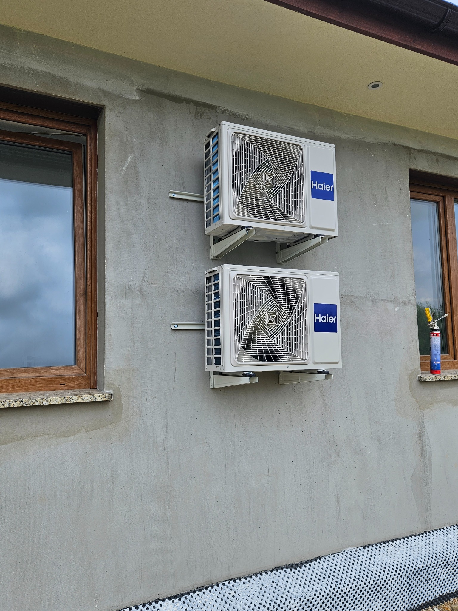 Dwa białe klimatyzatory Haier zamontowane jeden nad drugim na szarej, surowej ścianie budynku, widoczne okna z brązowymi ramami i pianka montażowa na parapecie.