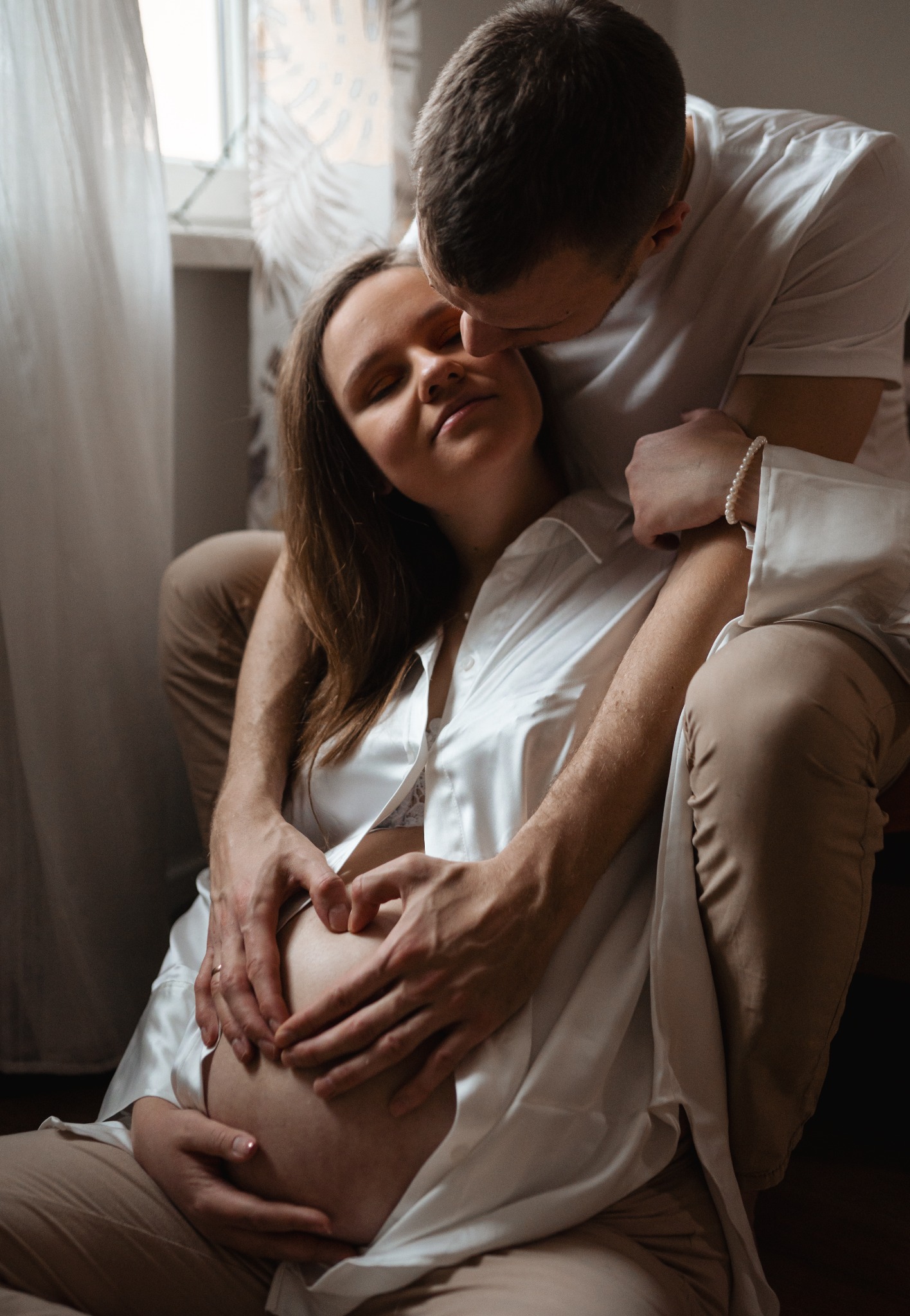 Przyszła mama w satynowej koszuli siedzi na podłodze, partner obejmuje ją, formując dłońmi serce na brzuchu, przytulając się do jej skroni.