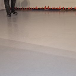 Epoxy3D - Świeżo wylana, błyszcząca posadzka żywiczna w szarym odcieniu, z widocznymi śladami po butach z kolcami i pomarańczowymi znacznikami poziomymi przy ścianie.