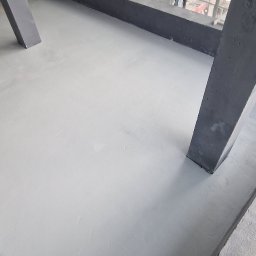 Epoxy3D - Świeżo wylana, jasnoszara posadzka w pomieszczeniu z betonowymi filarami i dużymi oknami, widok na plac budowy na zewnątrz.