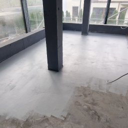 Epoxy3D - Wnętrze w trakcie prac wykończeniowych: częściowo pomalowana, szara posadzka w surowym pomieszczeniu z betonowymi filarami, widoczne okna i materiały budowlane w folii.