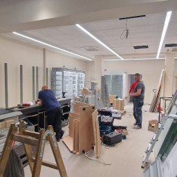 Epoxy3D - Montaż wyposażenia w nowym lokalu handlowym: pracownicy instalują lady chłodnicze i regały, widoczne narzędzia, drabina oraz materiały budowlane.