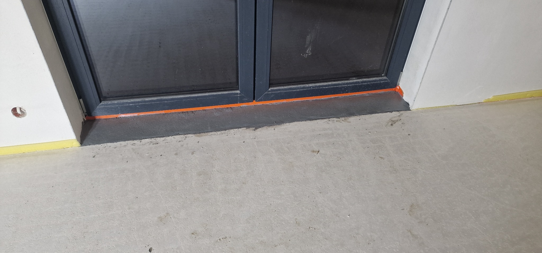 Szara, surowa posadzka betonowa przy oknie z czarną ramą, zabezpieczonym pomarańczową taśmą, żółta taśma malarska przy ścianie, widoczne ślady po pracach wykończeniowych.