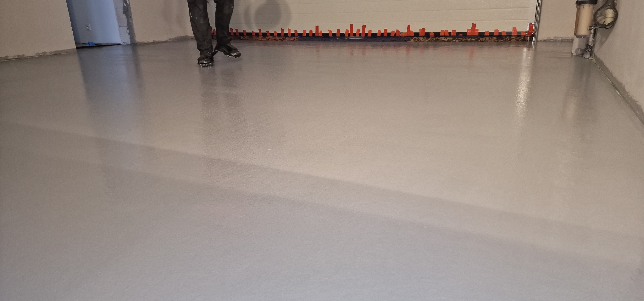 Świeżo wylana, błyszcząca posadzka żywiczna w szarym odcieniu, z widocznymi śladami po butach z kolcami i pomarańczowymi znacznikami poziomymi przy ścianie.