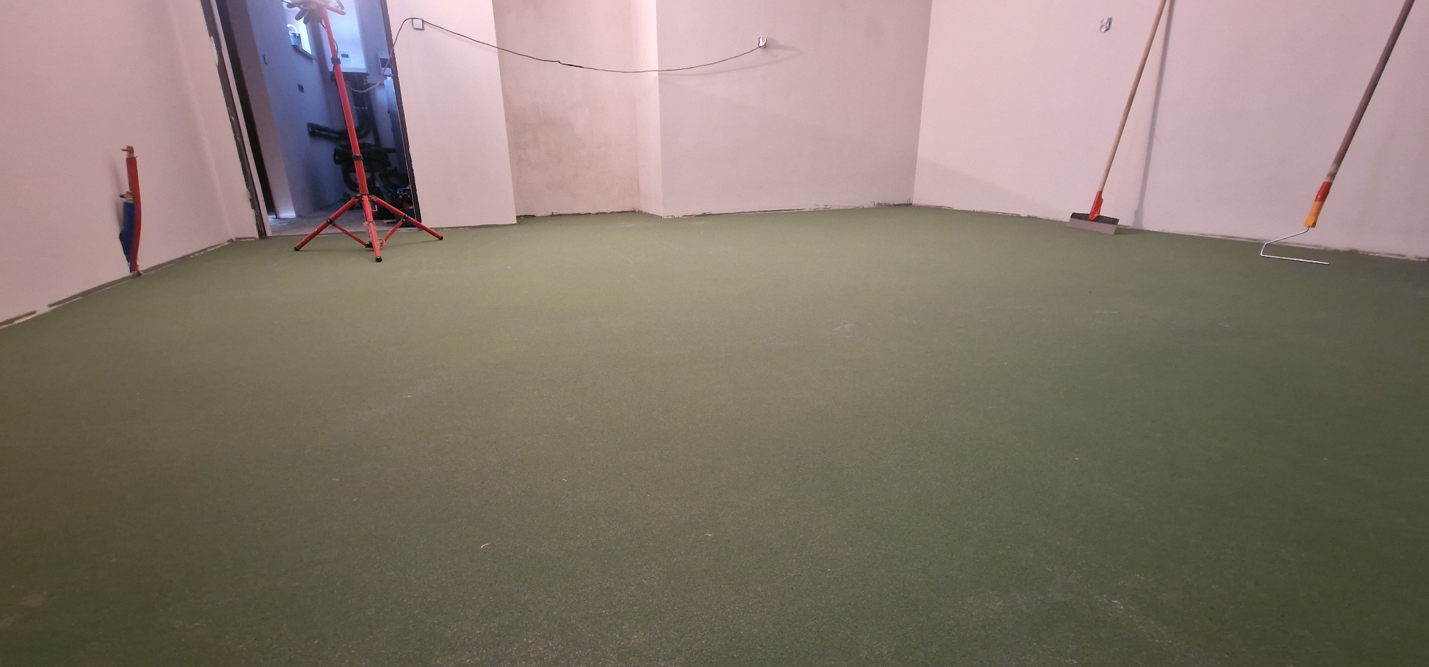 Świeżo wylana, zielona podłoga w pustym pomieszczeniu, widoczne narzędzia do rozprowadzania masy i lampa oświetleniowa.