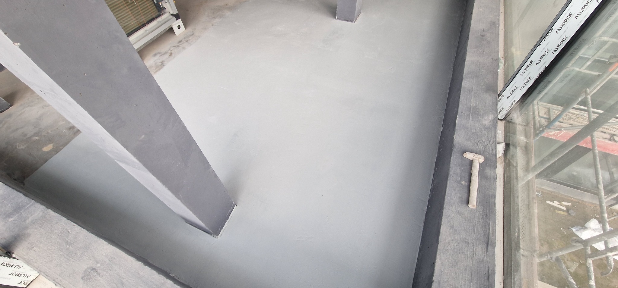 Świeżo wylana, jasnoszara podłoga, prawdopodobnie samopoziomująca w pomieszczeniu z widocznymi słupami konstrukcyjnymi i oknem. Na parapecie okiennym leży mały młotek.