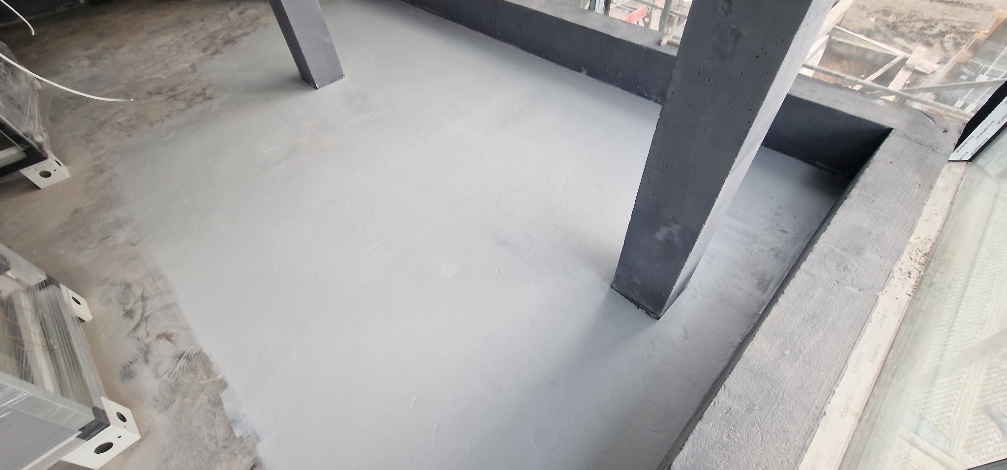 Świeżo wylana, jasnoszara posadzka w pomieszczeniu z betonowymi filarami i dużymi oknami, widok na plac budowy na zewnątrz.