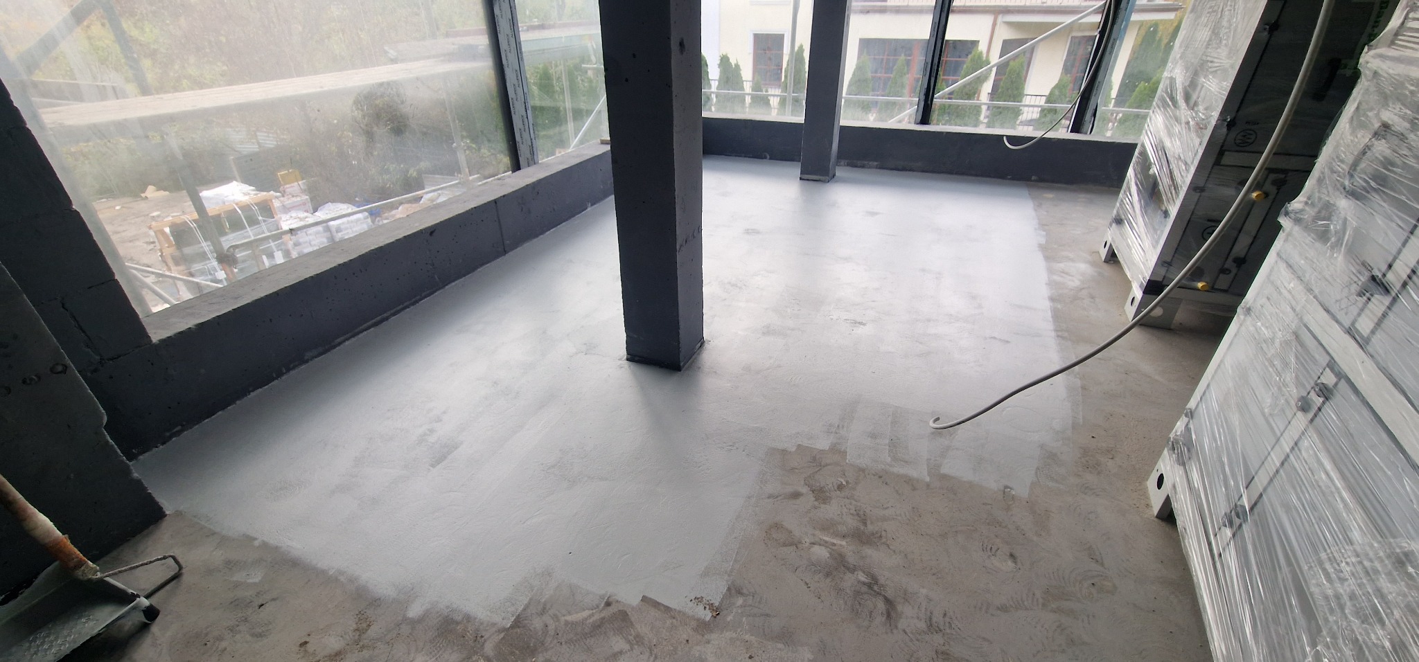 Wnętrze w trakcie prac wykończeniowych: częściowo pomalowana, szara posadzka w surowym pomieszczeniu z betonowymi filarami, widoczne okna i materiały budowlane w folii.