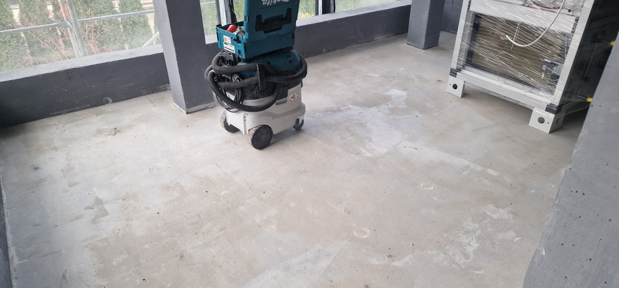 Przygotowanie podłoża pod posadzkę żywiczną: szlifowanie i odkurzanie betonu z użyciem przemysłowego odkurzacza Makita, widoczne ślady szlifowania i resztki pyłu. W tle okno i elementy...