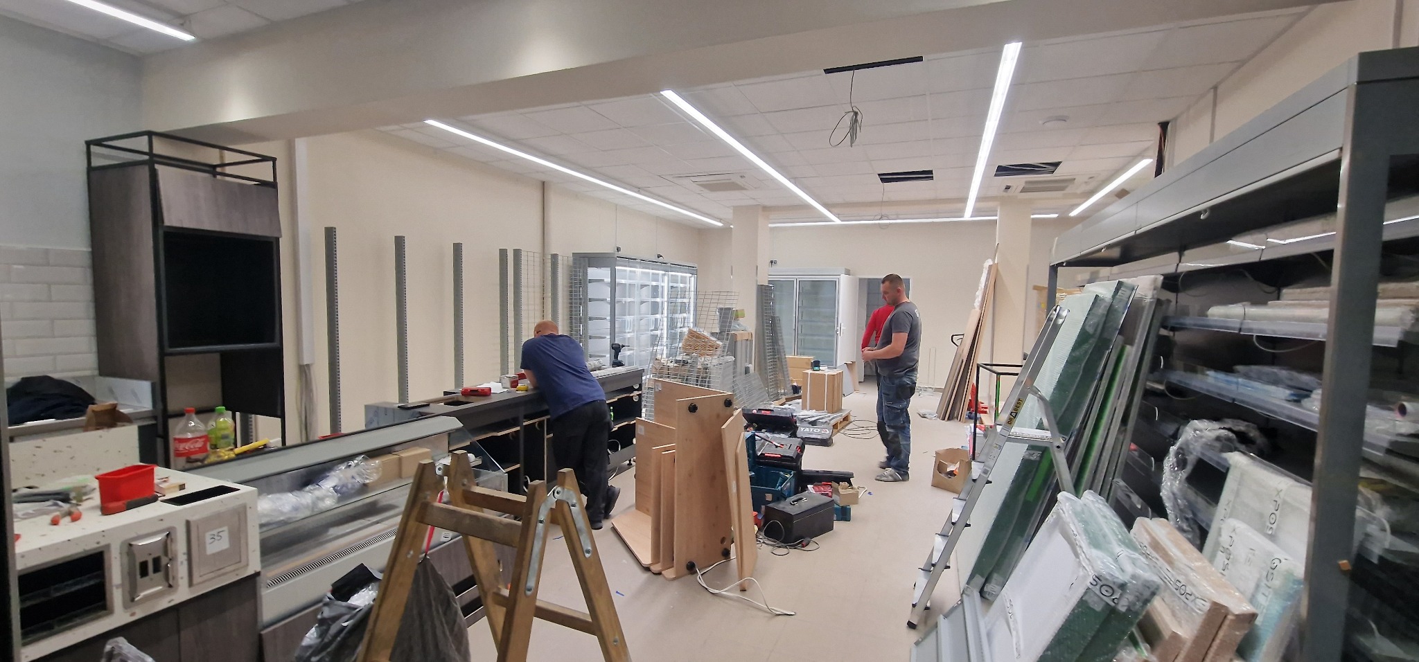 Montaż wyposażenia w nowym lokalu handlowym: pracownicy instalują lady chłodnicze i regały, widoczne narzędzia, drabina oraz materiały budowlane.