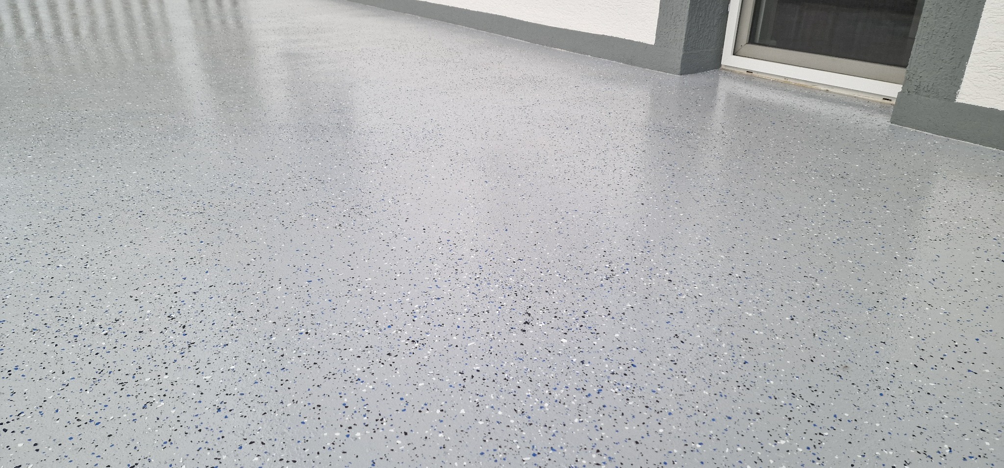 Jasnoszara posadzka z kolorowymi drobinkami, wykonana w Ciechanowie, o wysokim połysku, widoczna w narożniku pomieszczenia z oknem.