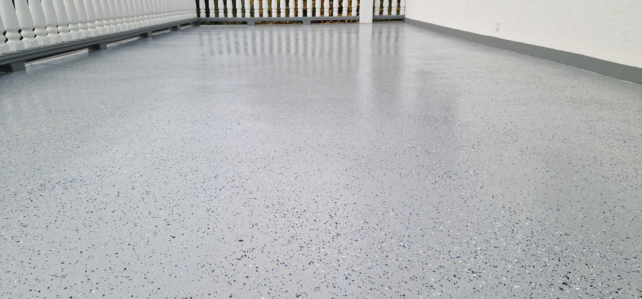 Jasnoszara, błyszcząca posadzka z drobnymi, kolorowymi wtrąceniami na balkonie, widoczna biała balustrada i fragment ściany.