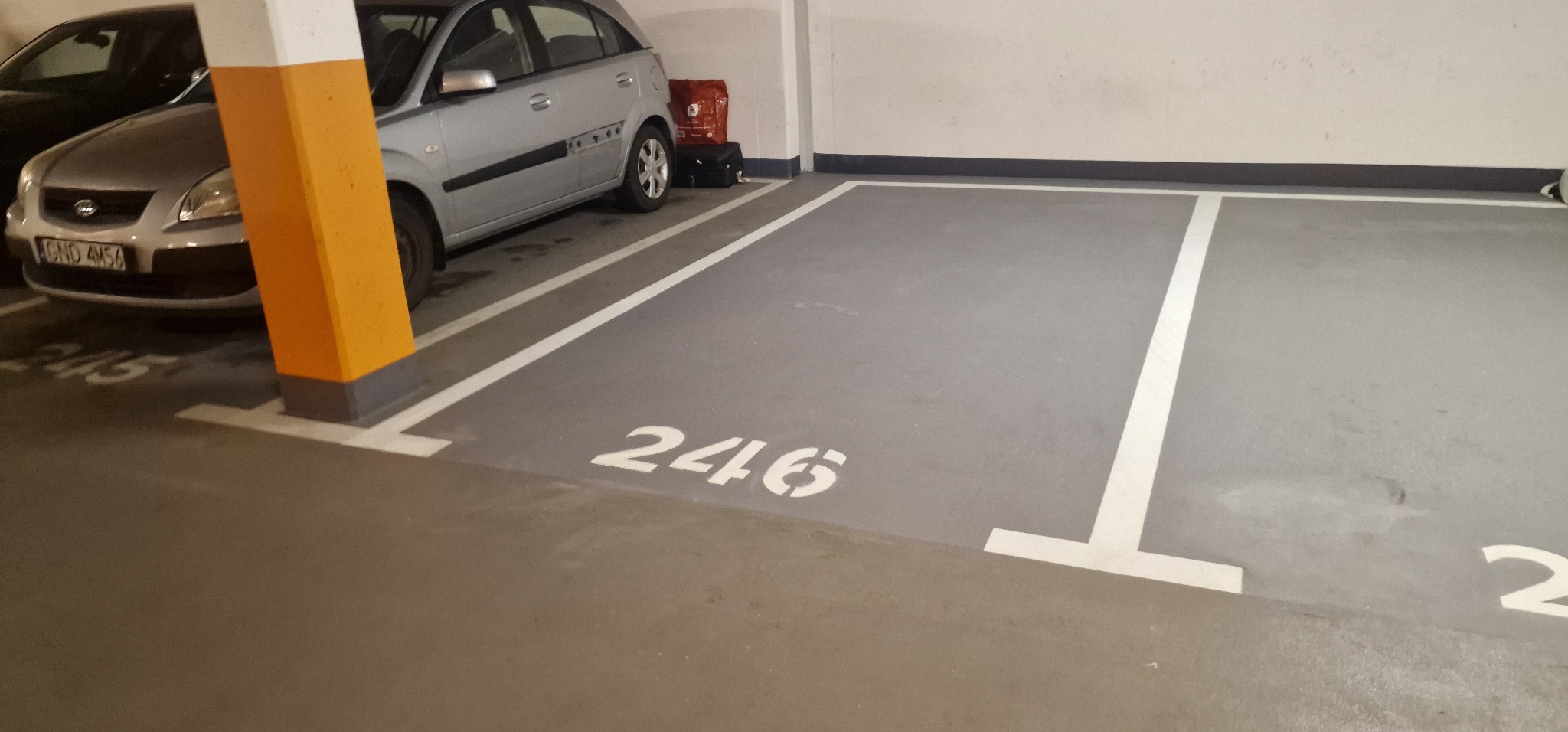 Szare miejsce parkingowe w garażu podziemnym z numerem 246, wyznaczonymi białymi liniami i pomarańczowym słupem, obok zaparkowany srebrny samochód.