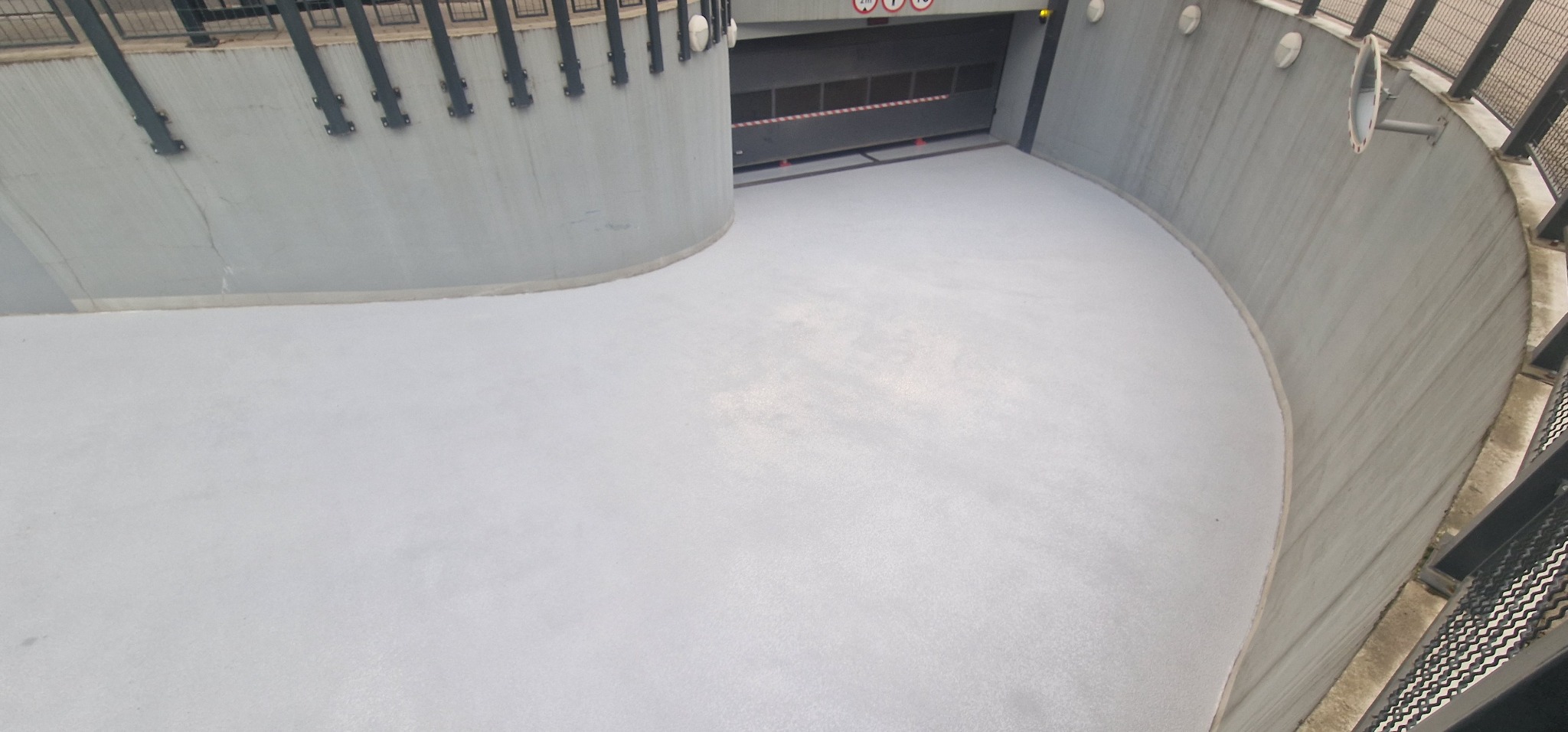 Gładka, jasnoszara posadzka wjazdowa do garażu podziemnego z szarymi ścianami wykończonymi metalowymi elementami.