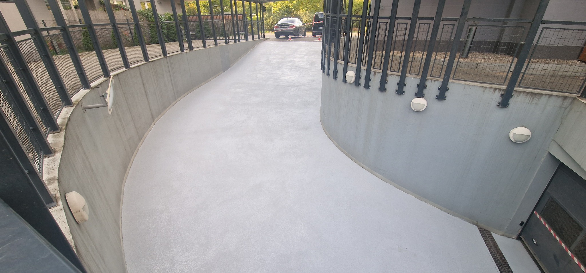 Biała, gładka posadzka wjazdowa do garażu podziemnego, z widocznymi lampami na ścianach i metalową barierką.