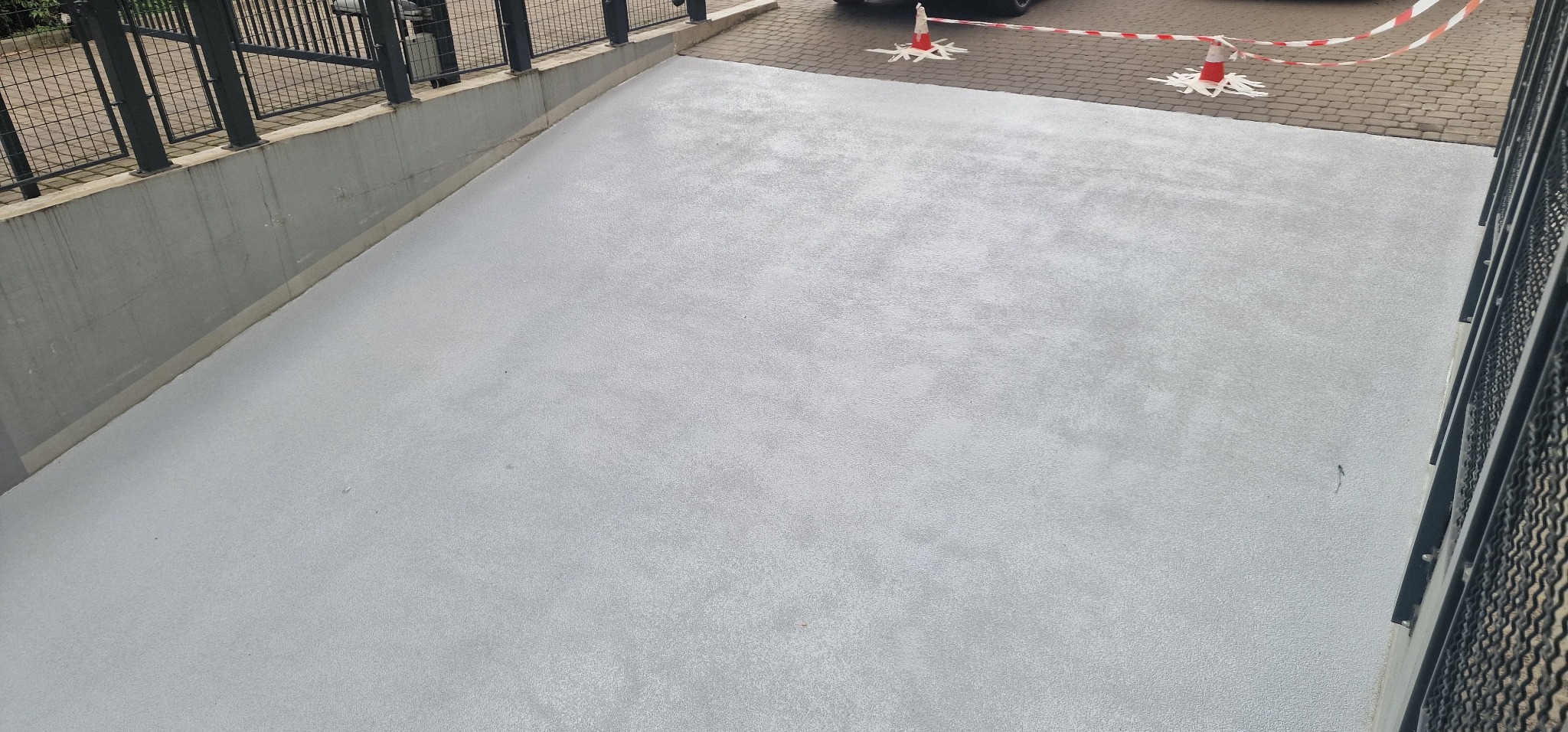 Świeżo wylana, jasnoszara powierzchnia z żywicy epoksydowej, zabezpieczona stożkami ostrzegawczymi i taśmą, widok z góry, z elementami metalowej balustrady.