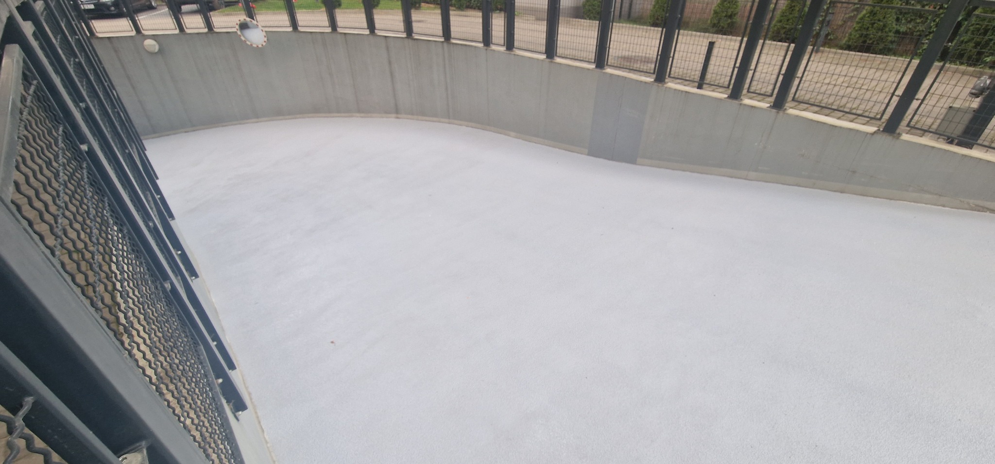 Jasnoszara, gładka powierzchnia posadzki w zagłębieniu, otoczona betonową ścianą i metalową balustradą, widok z góry.